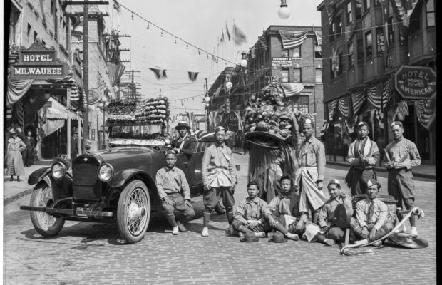 Black and white photograph of a group of dancers posing with a Lion Dance costume and musical instruments next to a car.