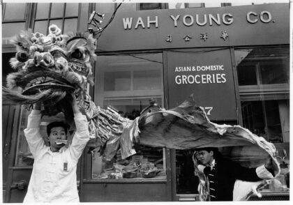 Black and white photograph of two dancers lifting a lion dance costume over their heads in front of Wah Young Co grocery store.