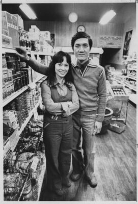 Black and white photograph of Thuy-Ngan and Hy-Van Nguyen standing inside their grocery store in front of shelves stocked with Vietnamese food products, books, and tapes.