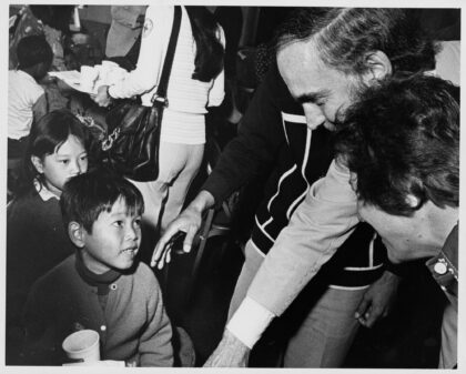 Black and white photograph of Washington Governor Dan Evans leaning down and smiling as he greets a young Vietnamese refugee child at Camp Murray.