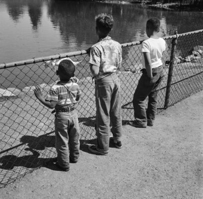 Boys standing on a beach wearing denim and holding fish.