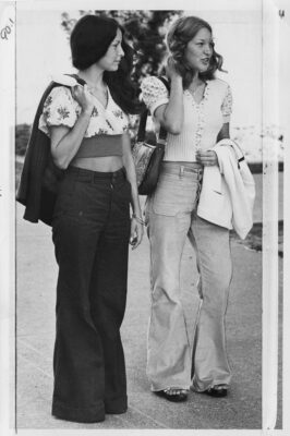 A black and white photo of two young women in denim bell-bottom pants and cropped tops, walking outdoors.