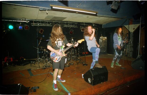A color photo of a rock band on stage, with a singer, guitarist, and bassist performing in a small venue wearing denim.
