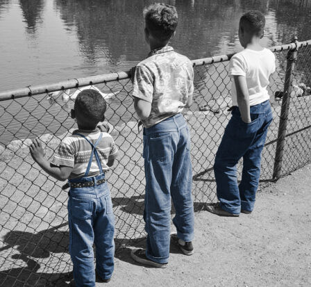 Boys standing on a beach wearing denim and holding fish.