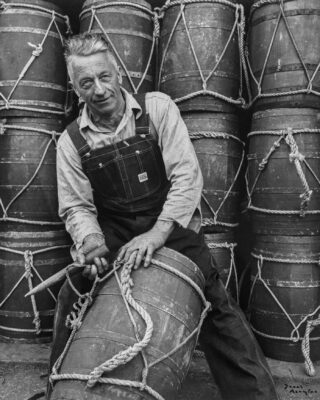 A black and white photograph of a fisherman wearing denim overalls in front of stacks of wooden barrels.
