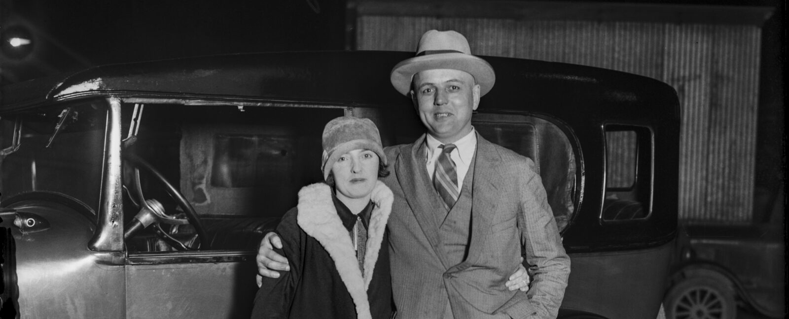 A historic, black and white photo of two people standing in front of a car.