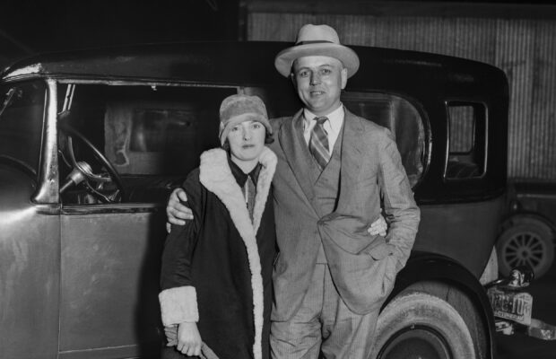 A historic, black and white photo of two people standing in front of a car.