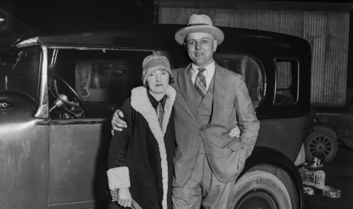 A historic, black and white photo of two people standing in front of a car.