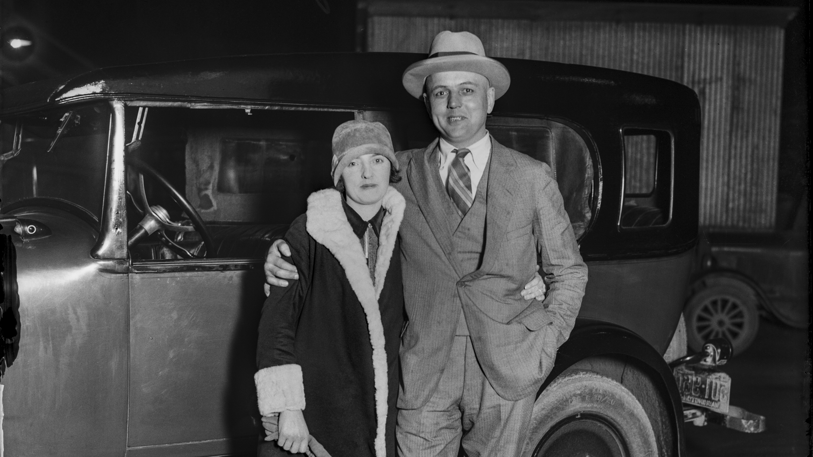 A historic, black and white photo of two people standing in front of a car.