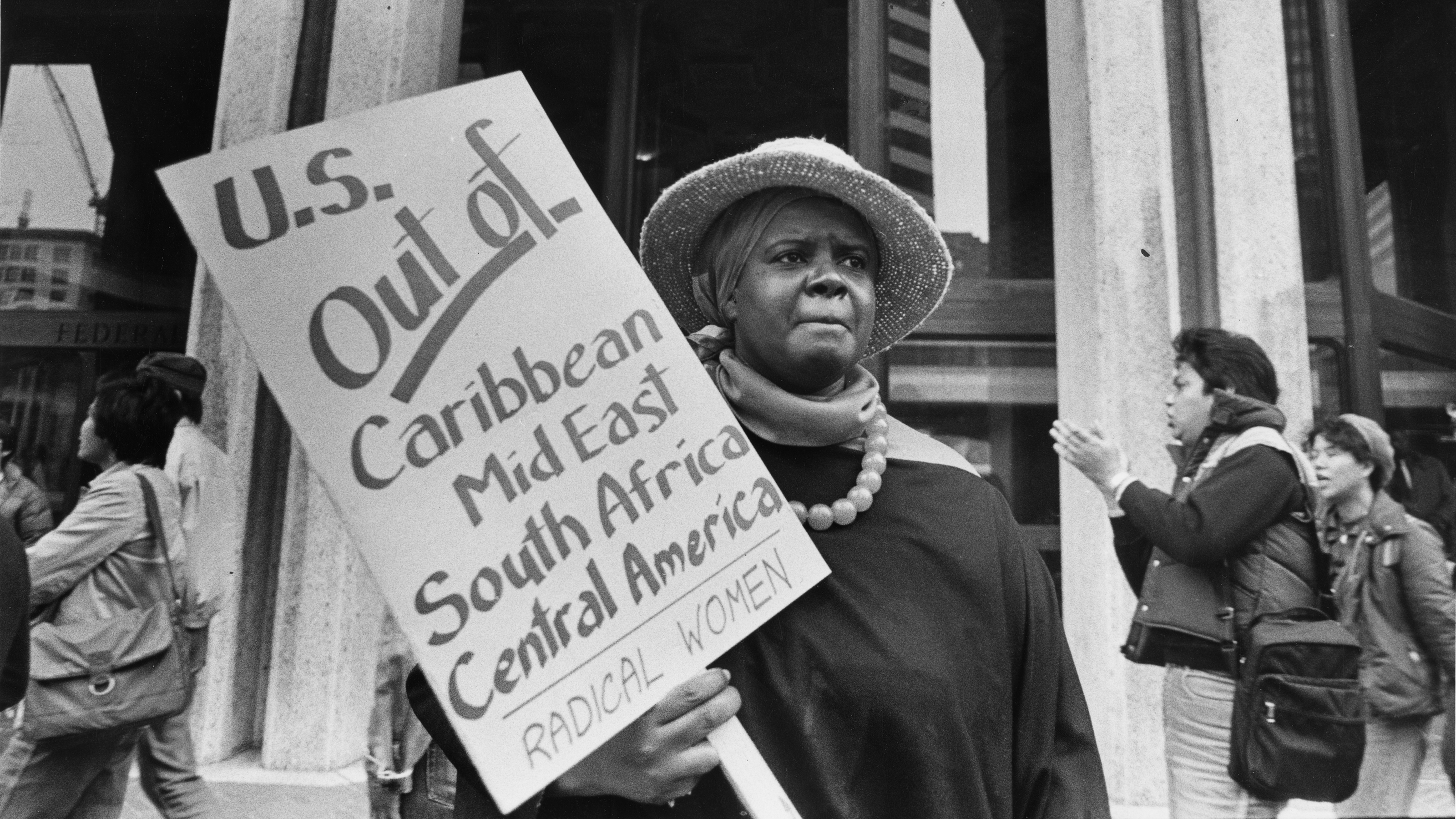 A woman holds a sign reading, "U.S. Out of Caribbean Mid East South Africa Central America Radical Women"