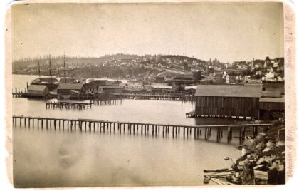 Black and white photograph of the Seattle waterfront with two long docks and several buildings.