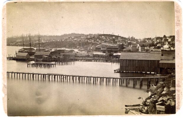 Black and white photograph of the Seattle waterfront with two long docks and several buildings.