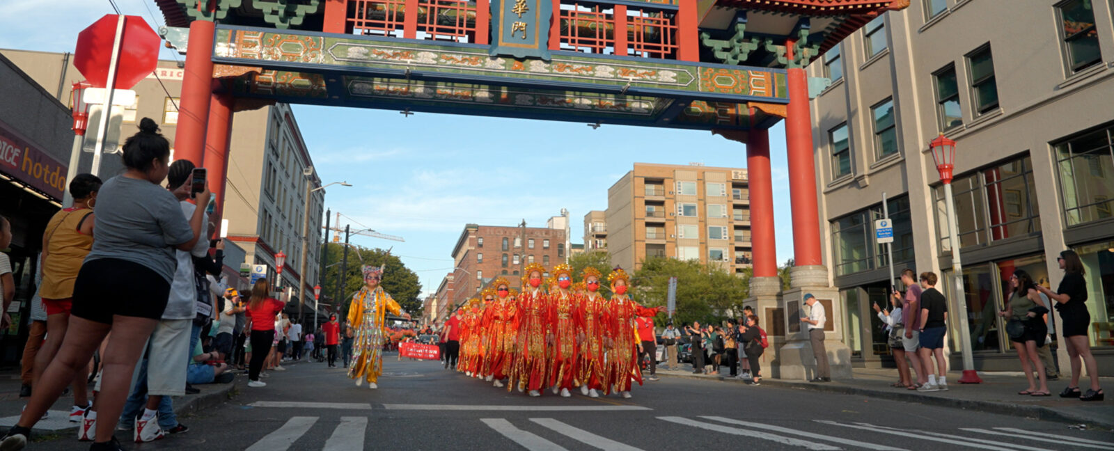 People marching in Seattle's International District beneath a Chinese paifang while spectators record video on their phones.