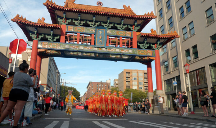 People marching in Seattle's International District beneath a Chinese paifang while spectators record video on their phones.