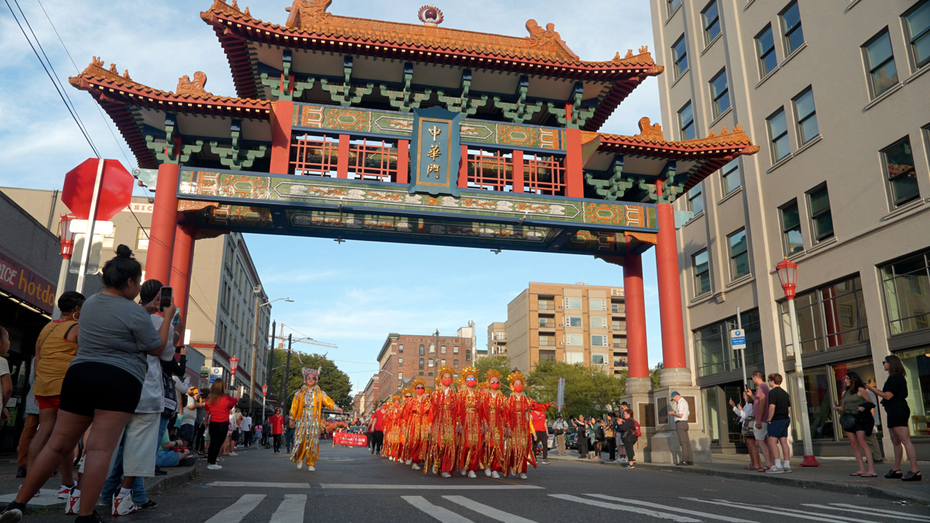 People marching in Seattle's International District beneath a Chinese paifang while spectators record video on their phones.