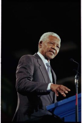 Nelson Mandela standing at a podium in front of a microphone wearing a dark colored suit.