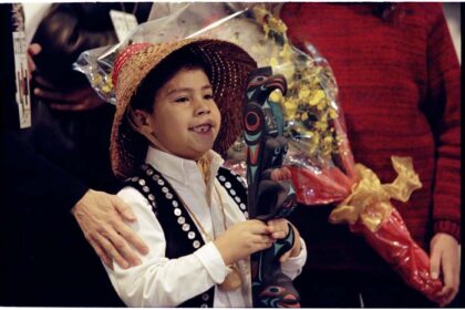 Dustin Williams holding a hand-carved Talking Stick wearing traditional Indigenous attire, with a smile on his face.