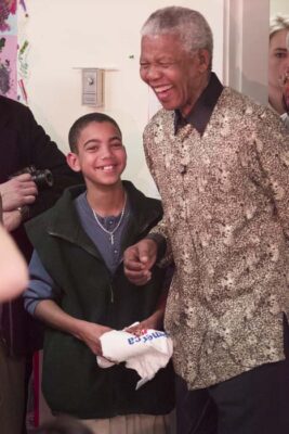 Nelson Mandela smiling and holding hands with a young boy during a joyful public appearance, surrounded by attendees and photographers.