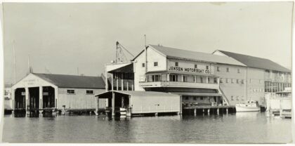 Black-and-white photo of the Jensen Motor Boat Company’s waterfront facility on Portage Bay, Seattle, showing the main building with large signage and several moored boats.