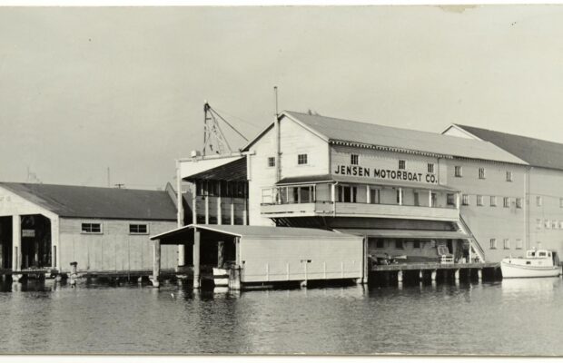 Black-and-white photo of the Jensen Motor Boat Company’s waterfront facility on Portage Bay, Seattle, showing the main building with large signage and several moored boats.