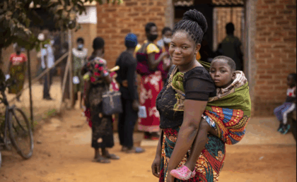 Mother carrying her child on her back while standing outside a rural health clinic in Africa, with other women and children waiting in the background.