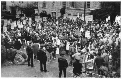 Large crowd of anti-apartheid demonstrators gathered outside the South African Consul’s residence in Seattle in 1985, holding protest signs and banners while police officers observe the peaceful protest.