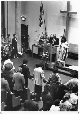 Rev. Dr. Samuel McKinney and Maryamu Eltayeb Givens lead a prayer at Mount Zion Baptist Church in Seattle during a 1990 celebration of Nelson Mandela’s release from prison, with attendees standing and holding hands in unity.