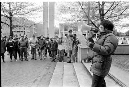 University of Washington students gather in Red Square in 1986 for an anti-apartheid protest, with one speaker using a megaphone and another holding a "Spend our $ wisely—Divest" sign to urge university divestment from South African investments.