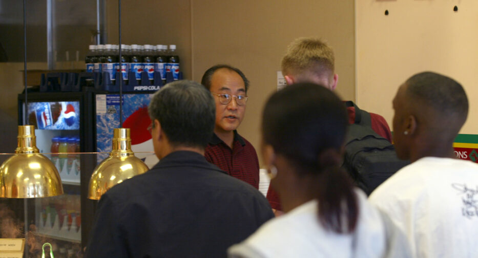 Jae Oh speaking to customers in line at Magic Garden Chinese Eatery, with beverages and condiments visible in the background.