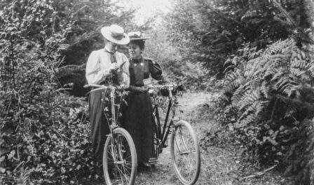 Two women with bicycles in the outdoors, August 24, 1898 surrounded by trees, ferns, and other Pacific Northwest foliage.