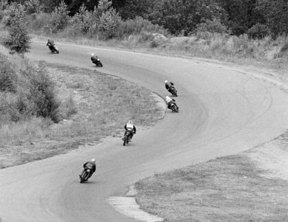 Motorcyclists racing around a curved track in Kent, Washington, during the All America Race Festival in 1966. Six riders lean into the turn on a tree-lined course surrounded by grass and brush.