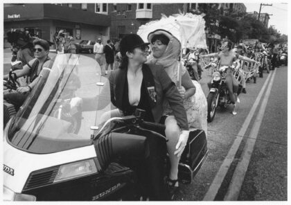 Two women riding a motorcycle with Dykes on Bikes at the Seattle Pride Parade in 1992. The lead rider wears a jacket and helmet, while the passenger holds a lace parasol, with other motorcyclists visible behind them.
