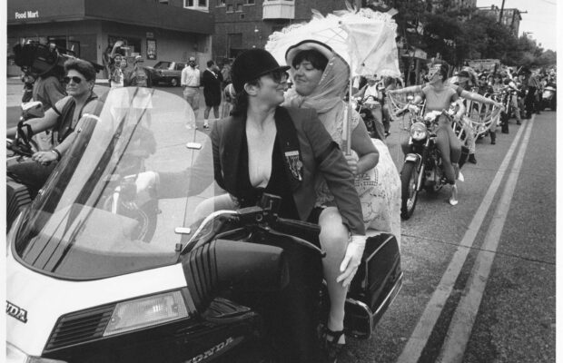 Two women riding a motorcycle with Dykes on Bikes at the Seattle Pride Parade in 1992. The lead rider wears a jacket and helmet, while the passenger holds a lace parasol, with other motorcyclists visible behind them.