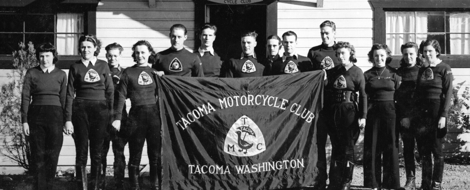 Fourteen male and female members of the Tacoma Motorcycle Club with a team banner in 1937.